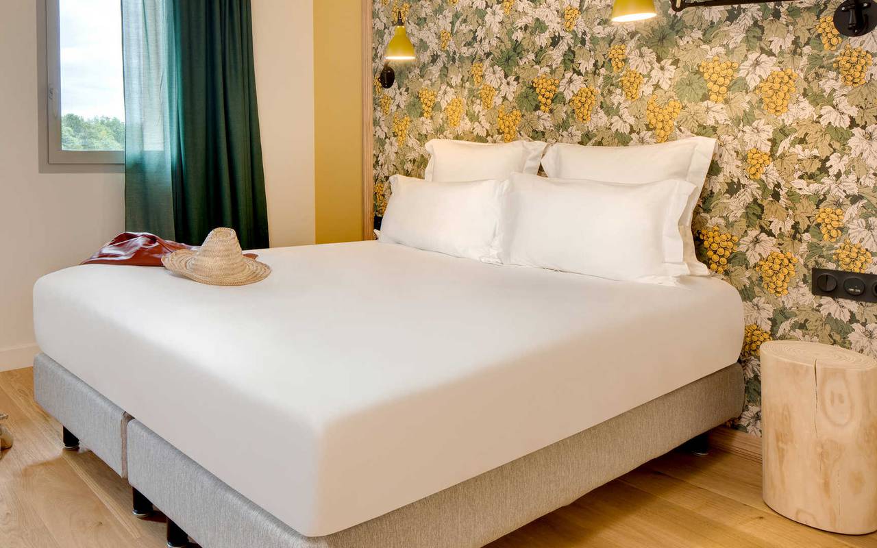 Vue d'un lit double dans une chambre aux murs jaunes, une fenêtre et un rideau vert dans un hôtel de charme à Saint Emilion. 
